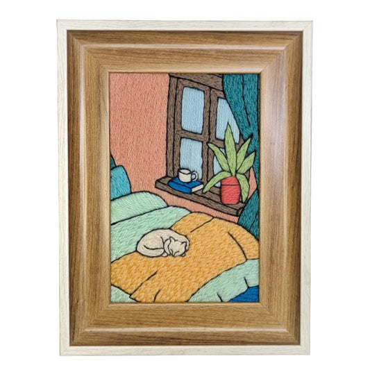 Framed artwork of a cat sleeping on a bed with a window and plant in the background.