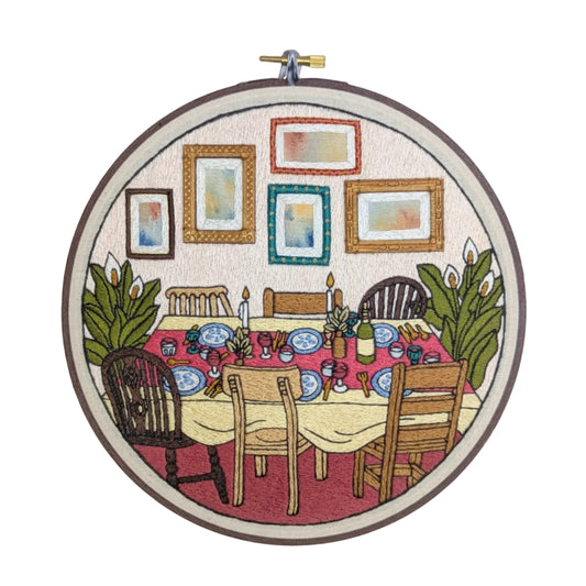 Embroidery hoop with a colorful depiction of a dining table and chairs, framed by pictures on a white background.
