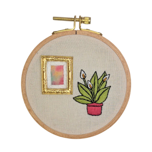Embroidery hoop with embroidered design of a plant and a small picture frame on beige fabric.
